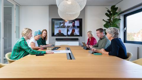 In de vergaderkamer zitten collega's in overleg en op het scherm zie je een aantal collega's die digitaal deelnemen aan de vergadering