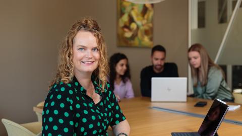 Een collega kijkt richting de camera terwijl de rest van het team op de achtergrond verder aan het vergaderen is