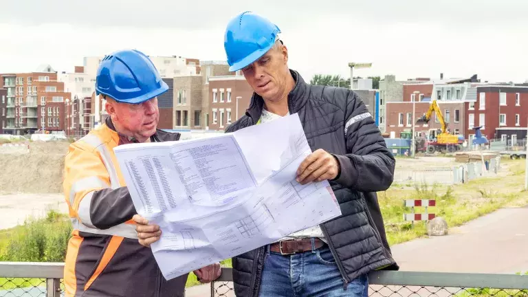 Twee collega's staan op de Melkwegbrug en kijken naar een groot vel met plannen. Op de achtergrond is de Weidevenne zichtbaar